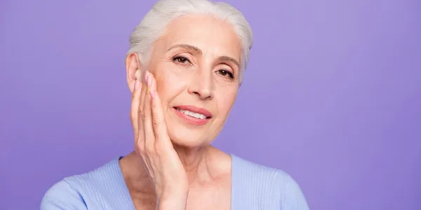 A woman with white hair is smiling and touching her face.